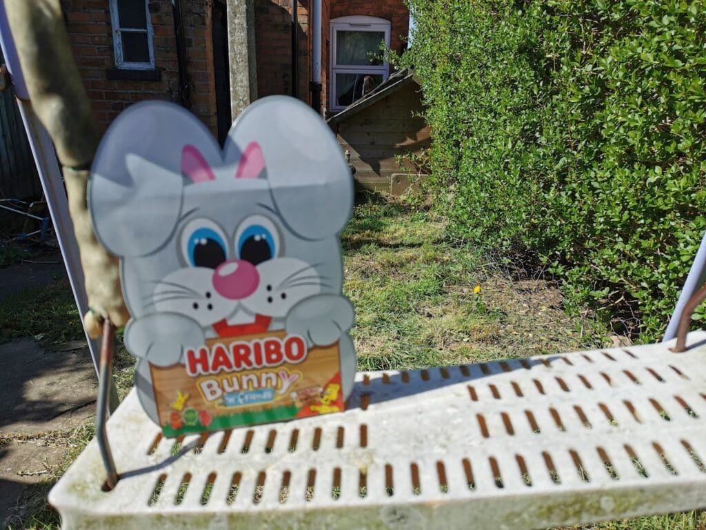 A Haribo Bunny cardboard cut-out is placed on a rusty white metal garden swing. Behind it, there is grass, a brick house, a shed, and a hedge on a sunny day. The bunny holds a box labelled "HARIBO Bunny.
