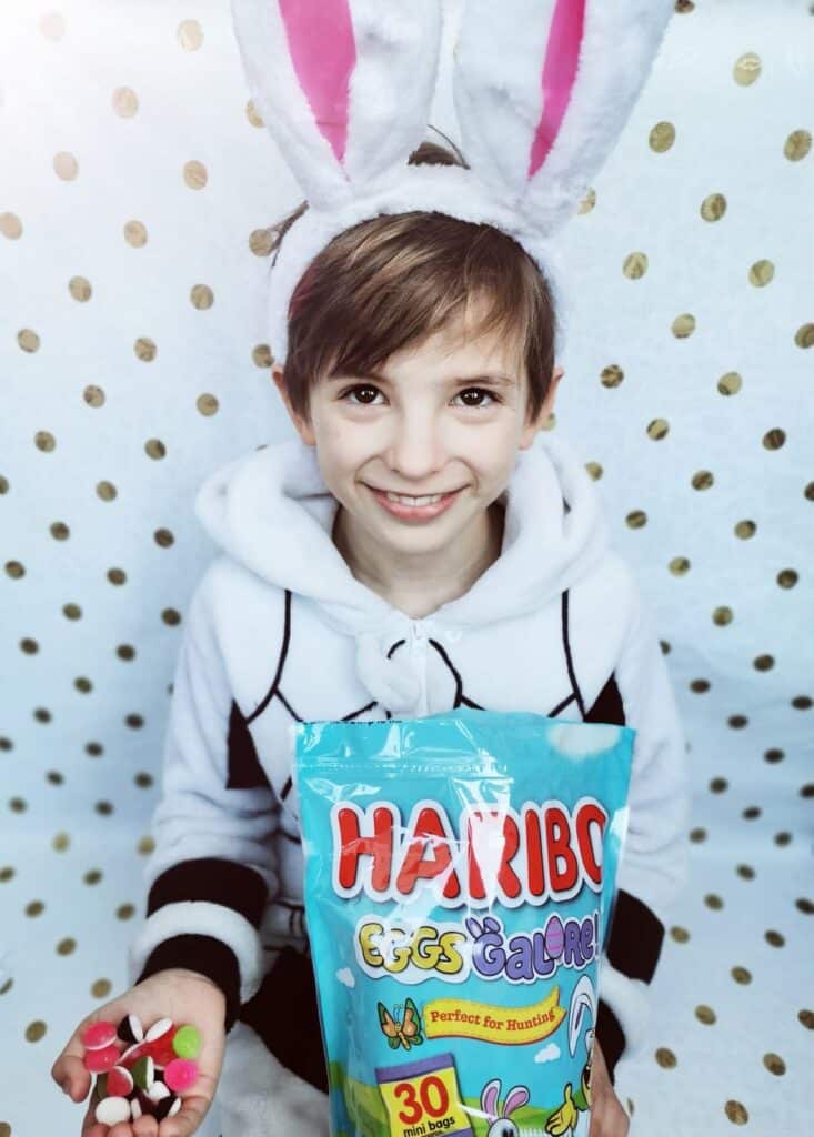 A smiling child wearing bunny ears and a white hoodie with black trim holds out colourful Haribo gummy sweets in one hand and a large Haribo Eggs Galore bag in the other, standing against a white backdrop with gold polka dots.