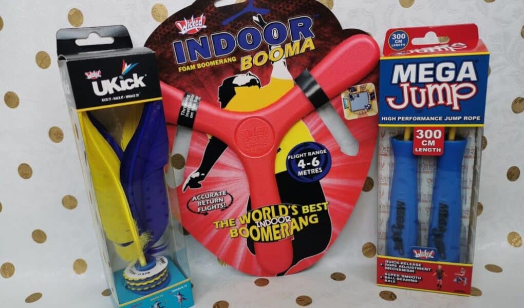 Three toys in packaging on a dotted background: a UKick shuttlecock (yellow, blue, black), a red foam Indoor Booma boomerang labelled “WORLD’S BEST INDOOR BOOMERANG,” and a blue Mega Jump high performance skipping rope, 300 cm length.
