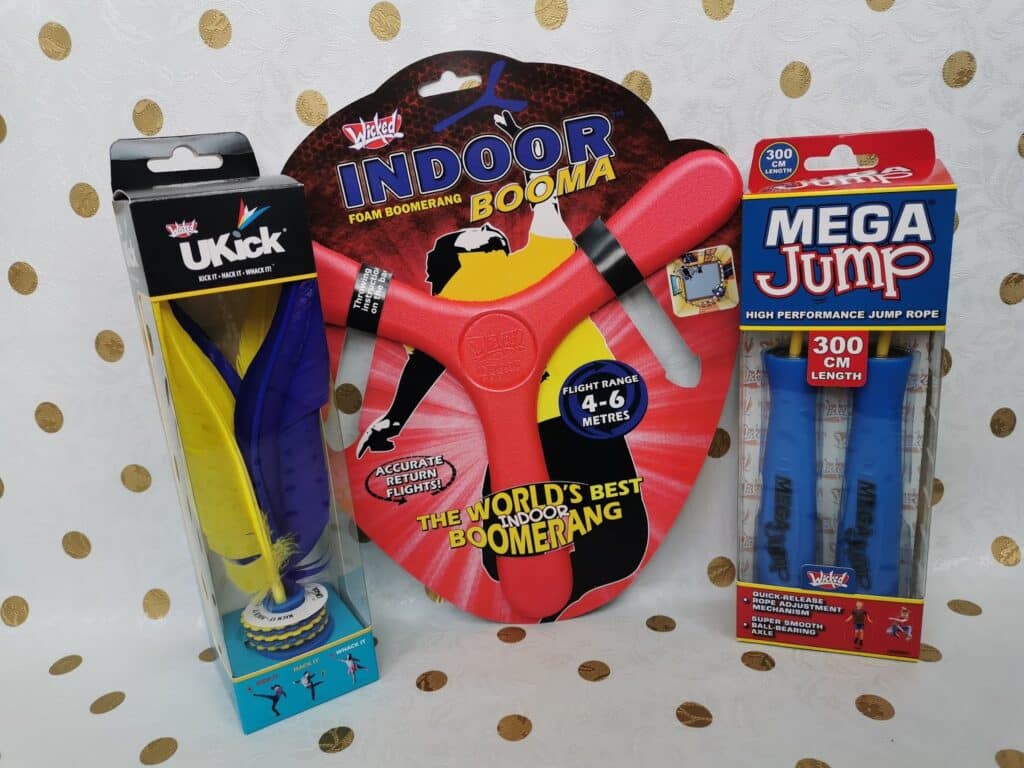 Three boxed toys on display: a yellow and blue UKick shuttle toy on the left, a red foam Indoor Booma boomerang in the centre, and a Mega Jump high-performance blue skipping rope (300 cm length) on the right, all set against a spotted background.