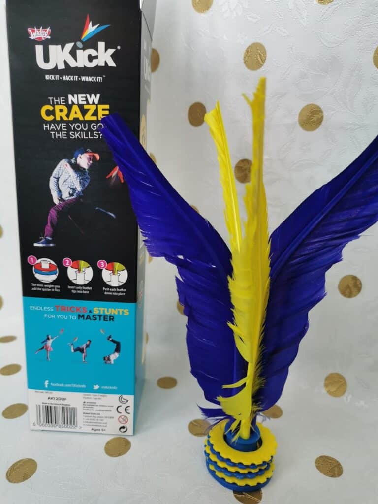 A toy called UKick stands upright, featuring blue and yellow feathers attached to a round base with alternating blue and yellow layers. The product box behind it displays images of people kicking the toy, brand logos, and colourful text describing tricks to master.