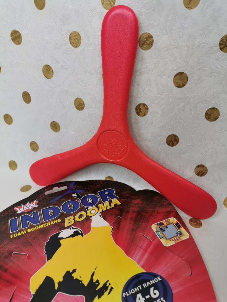 A red, three-pronged foam boomerang is propped against a white, textured wall with gold polka dots. Below it is a colourful product label reading "Wicked Indoor Booma," advertising a flight range of 4–6 metres.