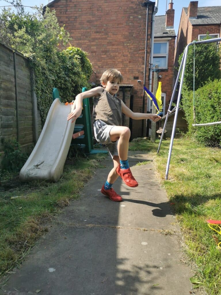 A young boy in shorts, red trainers, and a sleeveless top energetically poses with one leg raised and arms bent while holding a blue foam sword. He stands on a garden path beside a slide and swing set, with greenery and brick houses in the background.