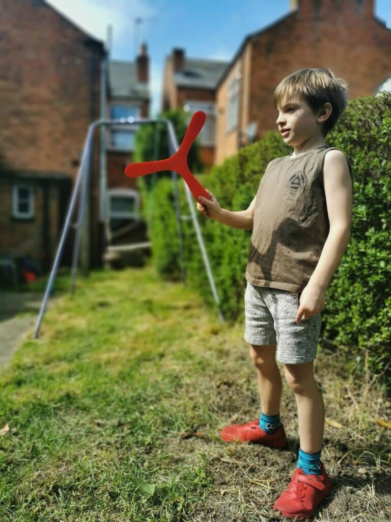 A young boy stands on grass in a back garden, holding a red boomerang. He wears a brown sleeveless vest, light shorts, blue socks, and red shoes. Behind him are bushes, a swing set, and brick houses under a sunny blue sky.
