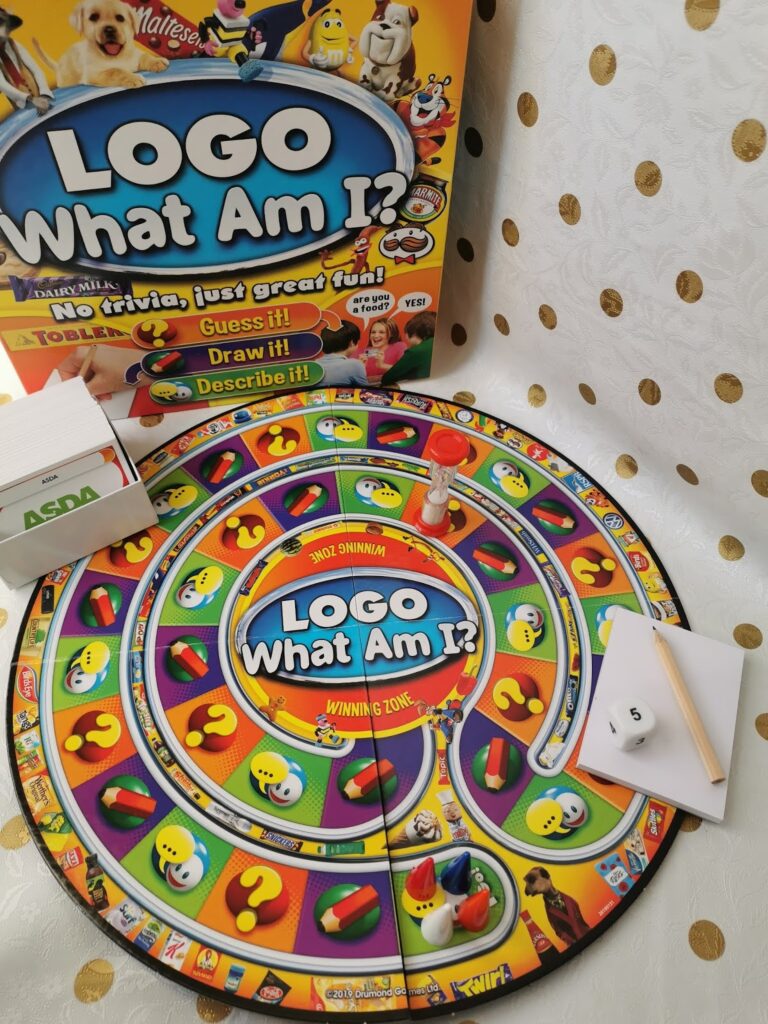 A colourful "LOGO What Am I?" board game is set up with the board, cards, box, pencil, and die. Game pieces are on the start. The circular board features question marks and emoji faces. The backdrop is a white fabric with gold polka dots.