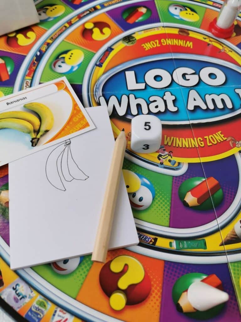 A colourful "LOGO What Am I?" board game is set up. Visible are a game card with a banana image, a hand-drawn banana sketch on paper, a pencil, a die showing 3 and 5, and part of the circular game board with icons and text.
