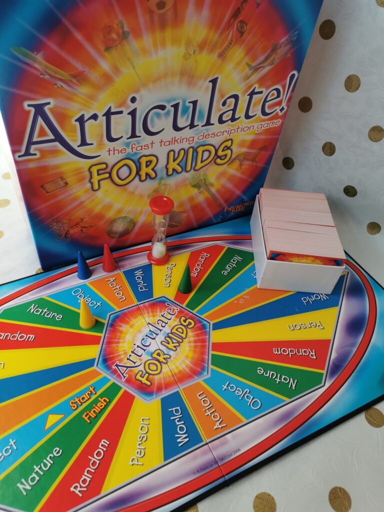A colourful board game called "Articulate! For Kids" is set up. The board shows segments labelled with topics like Nature, Person, World, and Object. A sand timer, cards, and game pieces are also visible next to the game box.