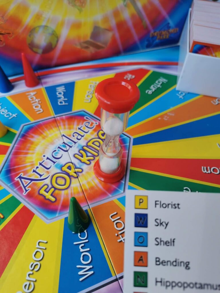 A close-up of the Articulate for Kids board game shows a red sand timer on the colourful board, game pieces, and a card listing words: Florist, Sky, Shelf, Bending, Hippopotamus. The board has rainbow sections with category names.