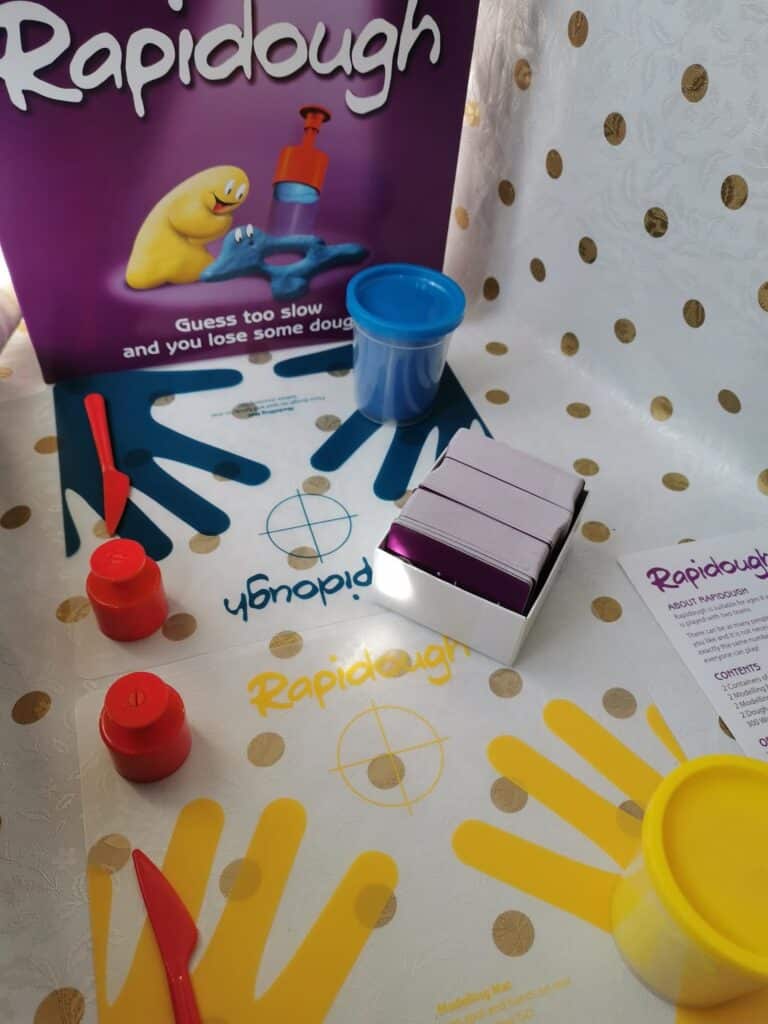 A Rapidough board game set-up on a gold polka-dot surface, showing game cards, red and yellow dough pots, a blue dough container, plastic tools, playing mats with handprints, and the purple game box featuring a yellow dough character.