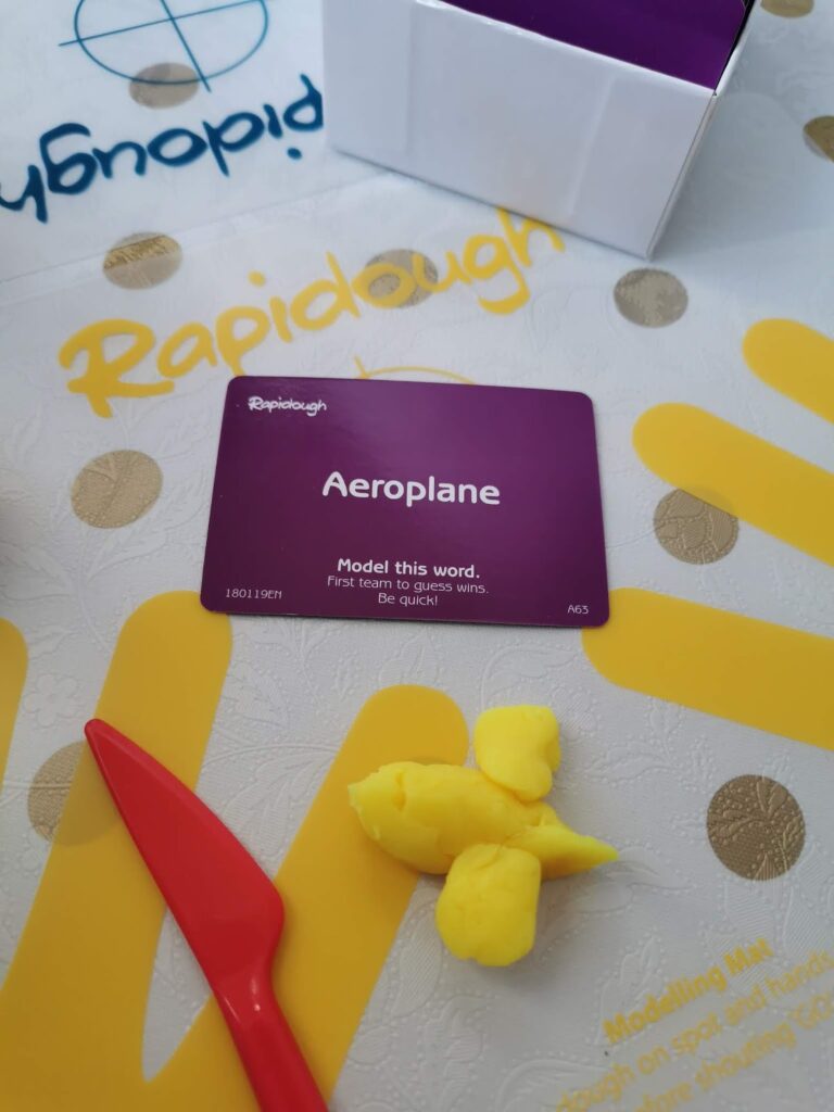 A purple Rapidough card with the word "Aeroplane" sits on a game mat. Below it is a red plastic modelling tool and a yellow dough model resembling an aeroplane. The mat shows part of the word "Rapidough" and yellow marks, with a box in the background.