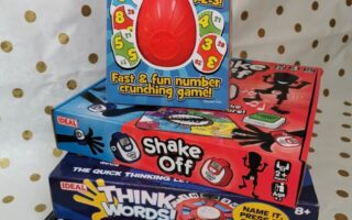 A stack of four colourful board games on a gold polka-dot background. The top game is “Tate 9” with a red egg-shaped piece, followed by “Shake Off,” “Think Words!,” and “Clock It!” Each box displays playful graphics and is by the brand Ideal.