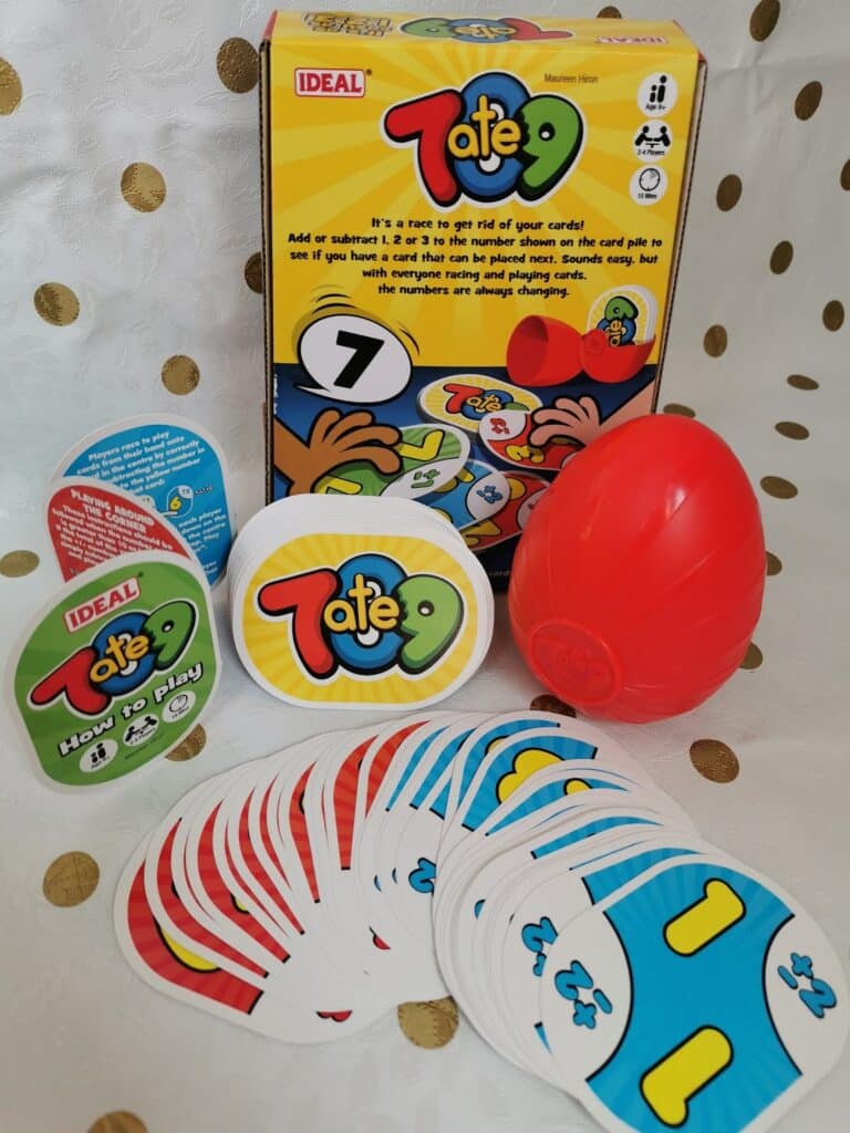 The image shows the Totep game set, including the game box, an egg-shaped red timer, rule cards, and several playing cards displaying colourful numbers and symbols, all laid out on a white tablecloth with gold polka dots.