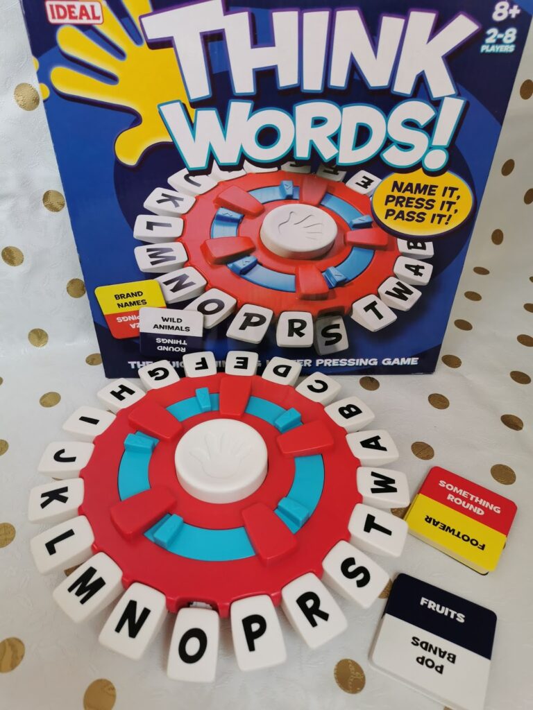 A board game called "Think Words!" is shown. The game piece has a blue and red circular base with white lettered tabs around the edge. The box stands behind, and several black cards with categories like "Wild Animals" and "Fruits" are visible.