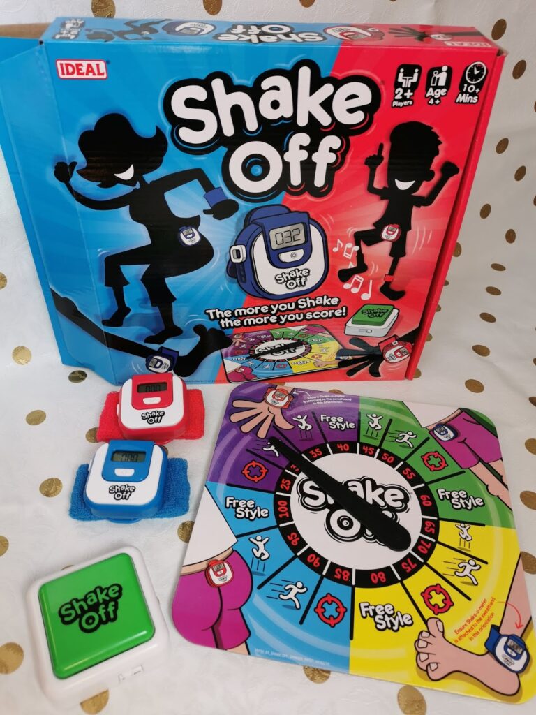 A Shake Off board game set includes a colourful game board, two blue and red wristbands with digital counters, a large green “Shake Off” button, and the game box featuring silhouettes of dancing figures and bold, playful graphics.