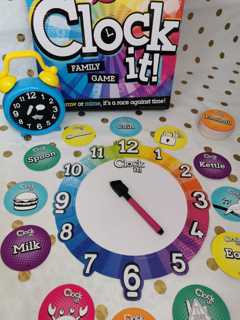 A colourful board game called "Clock it!" is displayed. It includes a round clock face with bold numbers, a pink dry-wipe marker, circular illustrated clue cards, and a small yellow toy clock, all on a white and gold spotty cloth.