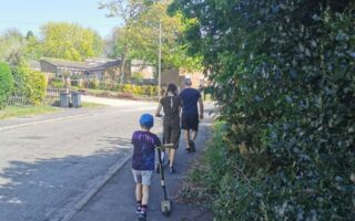 A boy wearing a cap and blue shirt pushes a scooter on a sunny pavement, walking behind two adults and a girl. Trees, bushes, and houses line the street, with shadows cast by the bright sunlight.