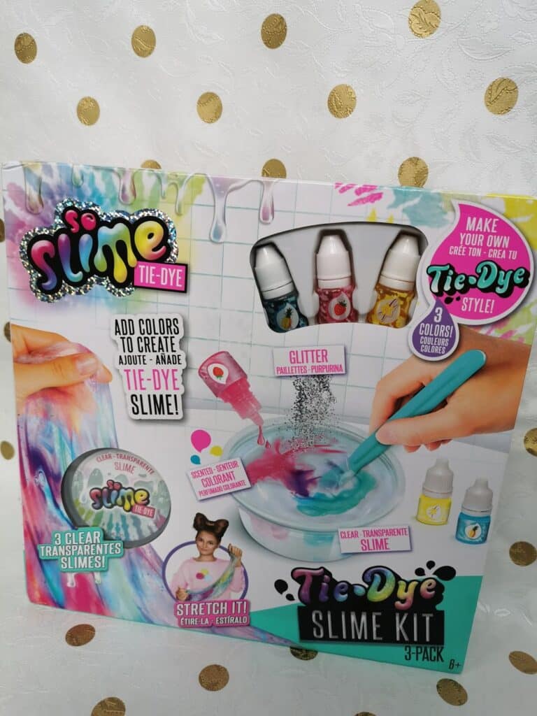 A colourful box for a So Slime Tie-Dye Slime Kit. The packaging shows bottles of colourant, glitter, a mixing tool, and clear slime. Text offers instructions and features, and the background has images of mixing and completed slime, with a pastel polka dot backdrop.