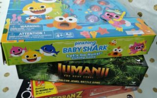 A stack of four board games is shown. On top is "Baby Shark Let's Go Hunt," featuring colourful cartoon sharks. Below are "Jumanji," "Hedbanz," and "Otrio" games. The background is white with gold polka dots.