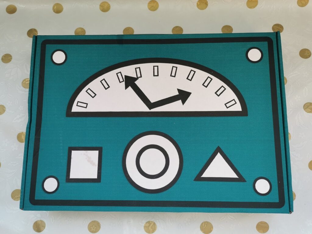 A teal rectangle with a graphic of a metre: a semicircular gauge with two black arrows pointing left and up, surrounded by tick marks. Below are three shapes: a square, a circle (with a central dot), and a triangle. Four white circles mark the corners.