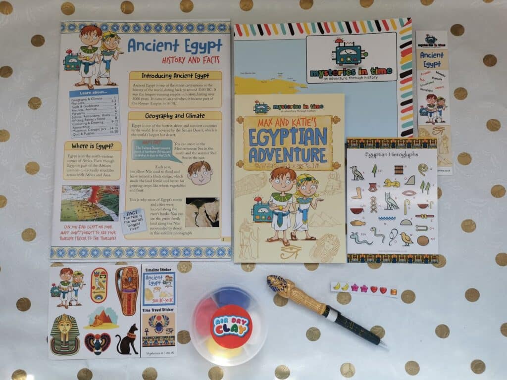 A collection of educational materials about Ancient Egypt on a gold polka dot background: fact sheets, storybooks, stickers, hieroglyphics chart, a pharaoh pen, modelling clay container, and small decorative gems arranged neatly.
