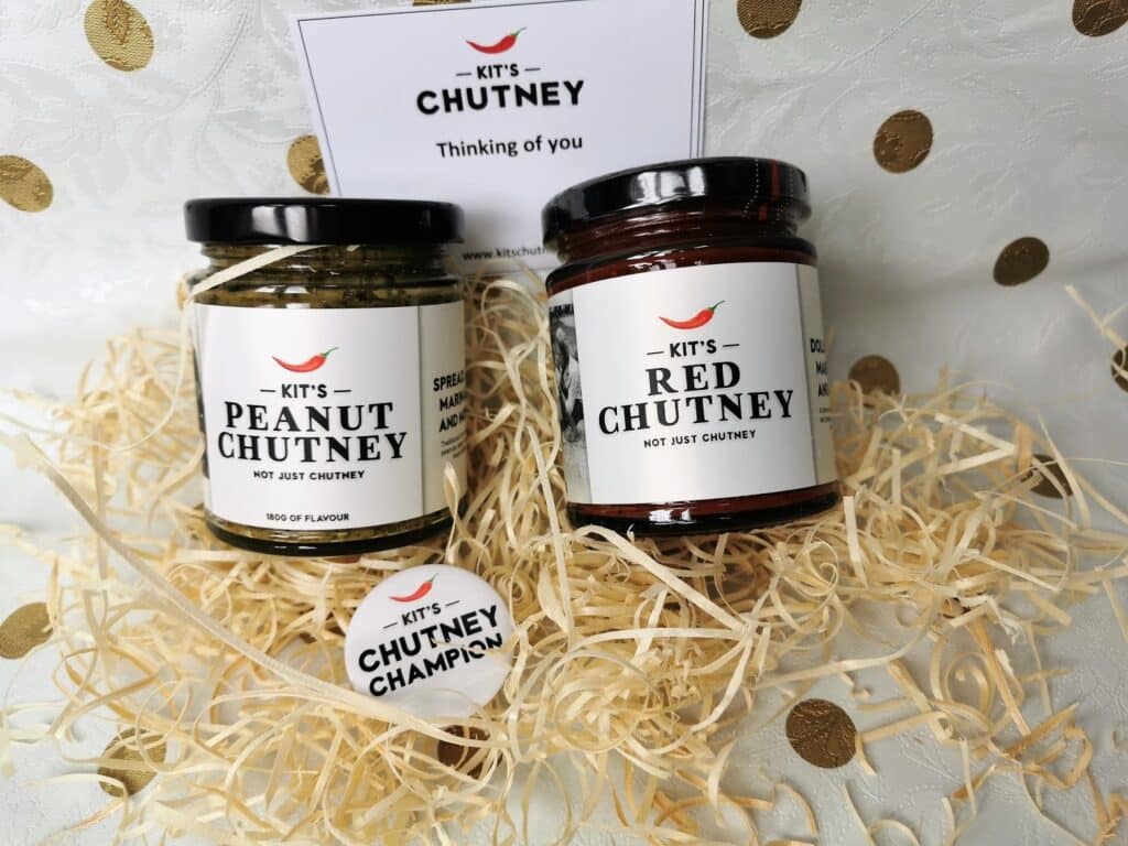 A gift box with shredded paper contains two jars labelled “Kit’s Peanut Chutney” and “Kit’s Red Chutney.” A round badge reads “CHUTNEY CHAMPION.” In the background is a card that says, “Kit’s Chutney—Thinking of you.” The setup is on a white, gold-dotted surface.