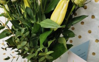 A glass vase holds a bouquet of white roses, unopened lilies, and green foliage. Next to the vase is a greeting card that reads "Hugs are good" in colourful letters. The background is white with a gold polka dot pattern.