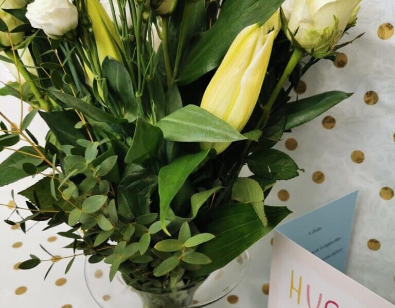 A glass vase holds a bouquet of white roses, unopened lilies, and green foliage. Next to the vase is a greeting card that reads "Hugs are good" in colourful letters. The background is white with a gold polka dot pattern.