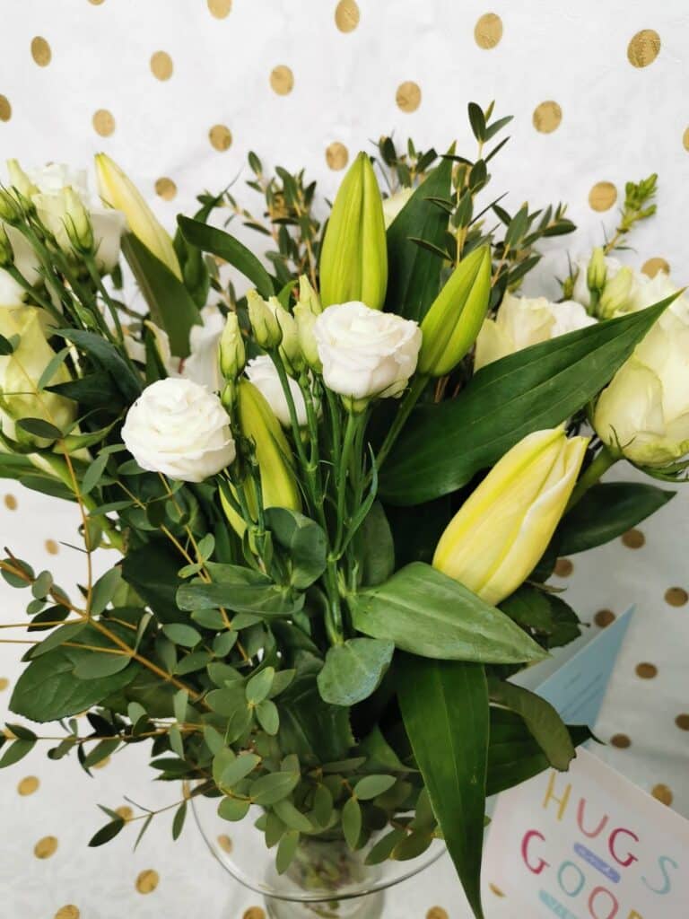 A bouquet of white roses, closed lily buds, and green foliage in a glass vase. The background features white paper with gold polka dots, and a partial greeting card with colourful letters is visible at the bottom right.