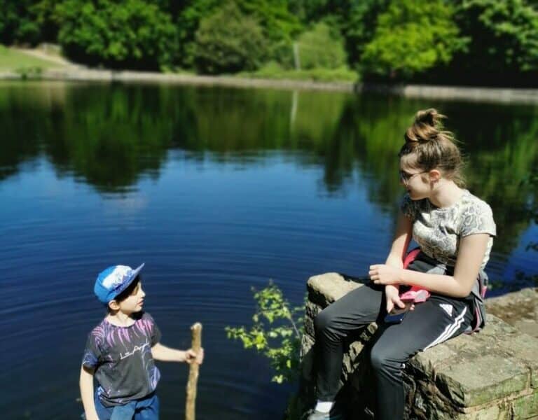 Two children are by a lake on a sunny day. A boy in a blue cap, holding a stick, stands at the water’s edge. A girl in a ponytail, patterned shirt, and trainers sits on a stone wall, looking at him. Green trees and a clear blue sky are in the background.