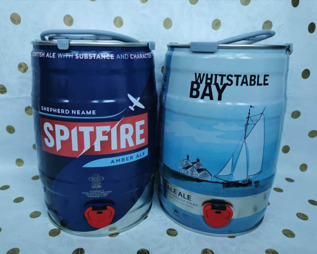 Two mini kegs of ale stand side by side on a white surface with gold polka dots. The left keg is dark blue with "Spitfire Amber Ale" branding; the right keg is light blue, labelled "Whitstable Bay Pale Ale," and features a coastal scene with a sailboat.