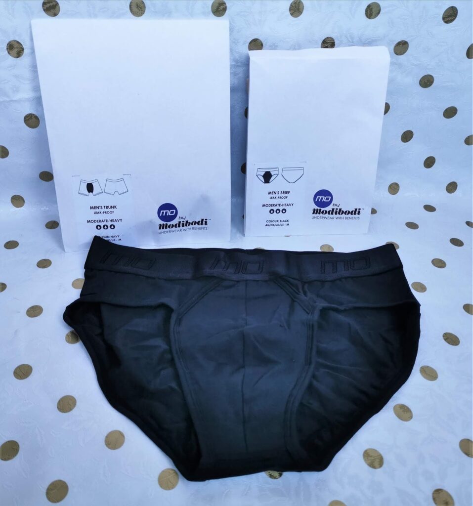 A pair of black men’s briefs is displayed in front of two white Modibodi packaging bags labelled “Men’s Trunk” and “Men’s Brief.” The background is white with gold polka dots. The briefs have a subtle waistband logo detail.