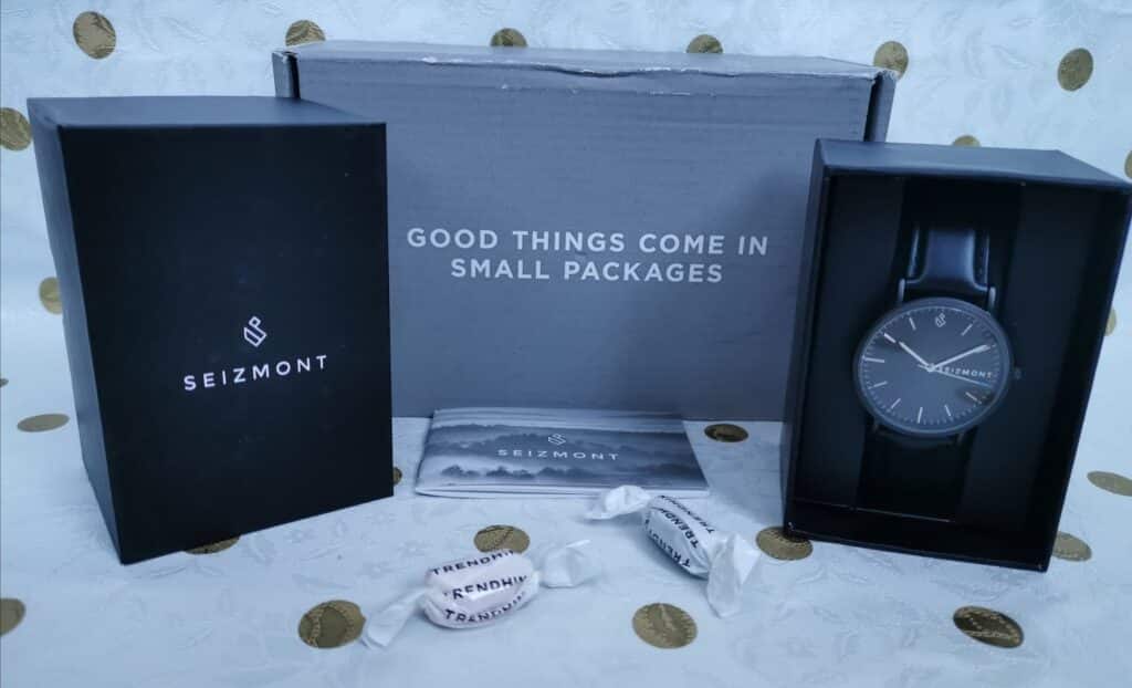 A Seizmont wristwatch with a black strap is displayed in an open black box on the right. To the left is a closed black Seizmont box. Behind them is a grey box with the text “GOOD THINGS COME IN SMALL PACKAGES.” Two sweets and a card are in front.