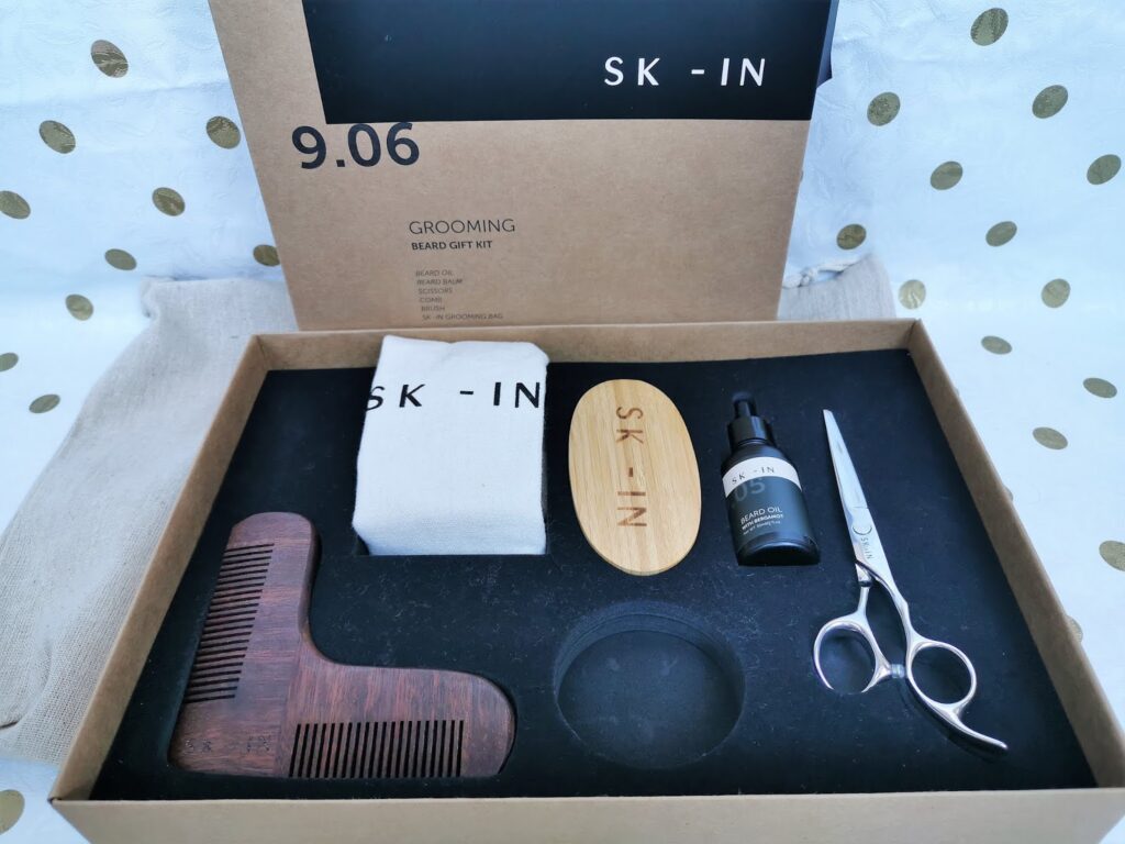 A men's grooming beard gift kit in a box includes a wooden beard comb, oval brush marked “SK-IN”, silver scissors, beard oil, a cloth bag, and a shaping tool. The packaging is minimalist, with brown and black colours and the brand “SK-IN” displayed.