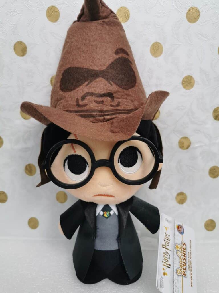 A plush doll of Harry Potter wearing large round glasses, a black robe, and a felt Sorting Hat with a face on it. The doll has a red lightning bolt scar on its forehead and a white shirt with a tie. The background is white with gold polka dots.