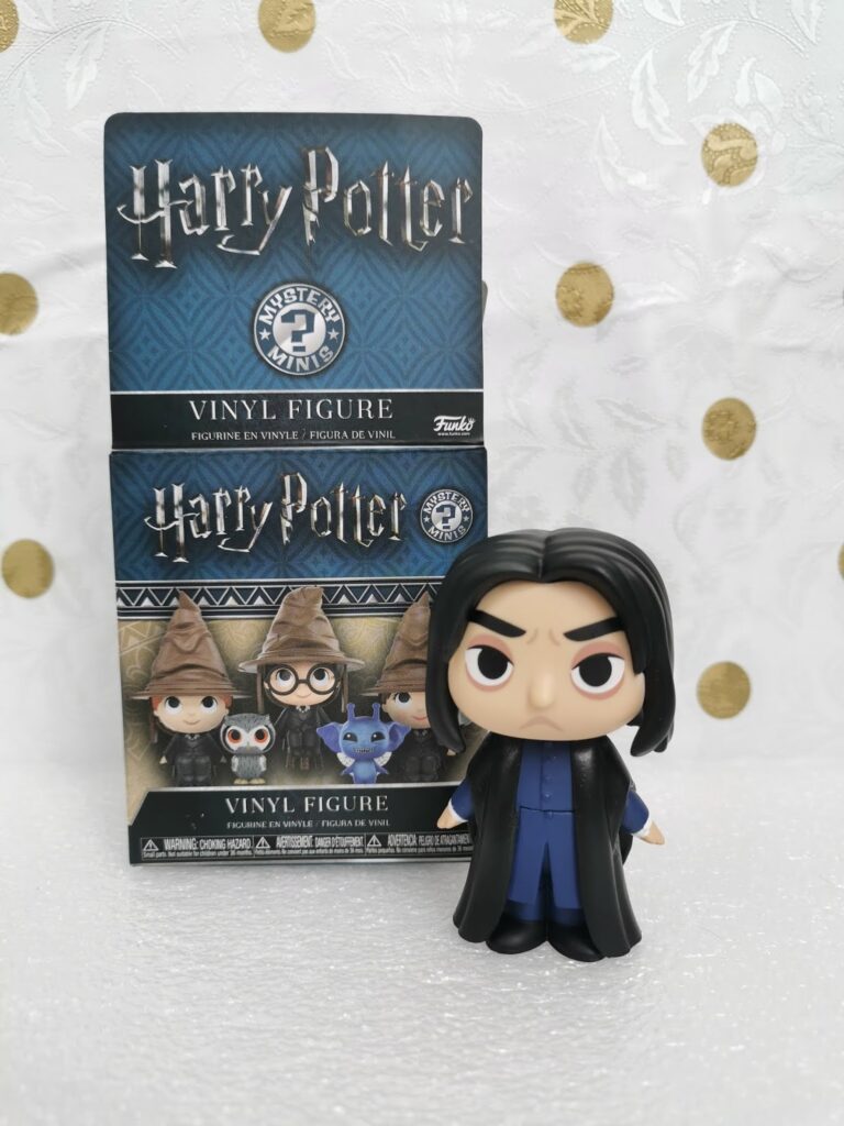 A Funko Mystery Minis vinyl figure of Severus Snape stands in front of its Harry Potter-themed box. The box features illustrations of other Harry Potter characters, the Sorting Hat, and "Harry Potter" in large gold letters. The background is white with gold dots.