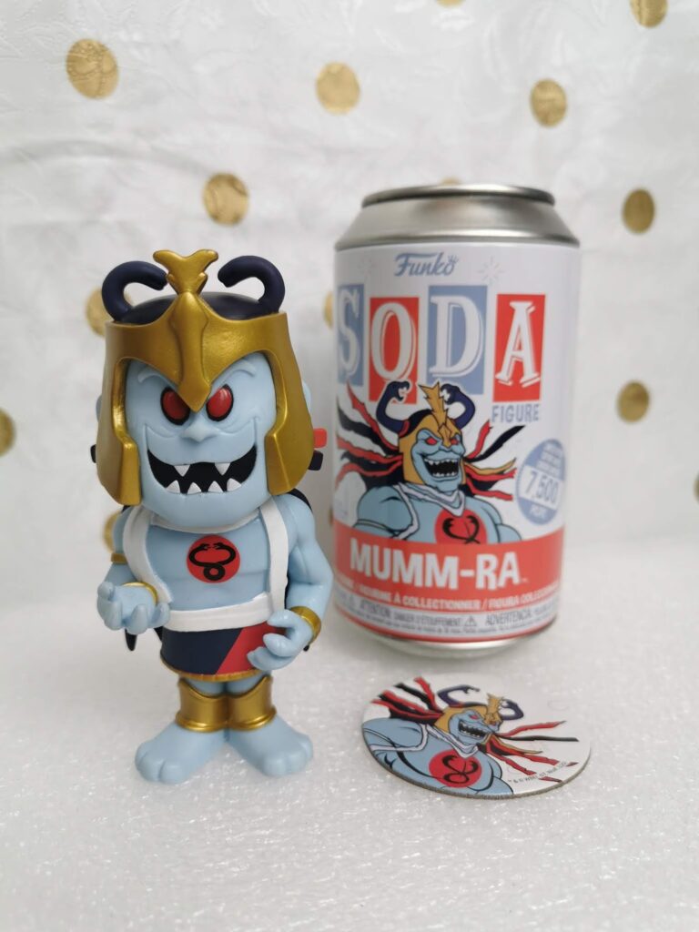 A vinyl figure of Mumm-Ra from ThunderCats, wearing gold armour and a helmet, stands in front of a Funko SODA can labelled "Mumm-Ra." A circular collector’s card with matching art is placed on a white surface with gold polka dots in the background.