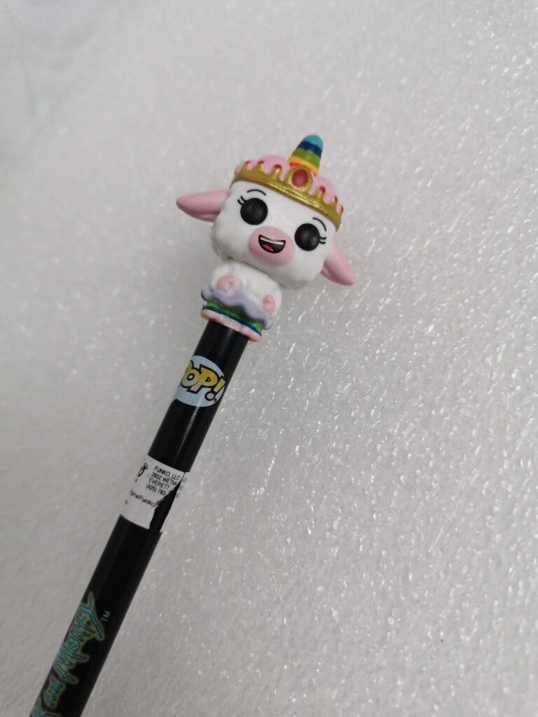 A black Funko Pop! pencil with a topper shaped like a smiling, white unicorn character with a rainbow horn, pink ears, golden crown, and small hooves, photographed against a textured white background.