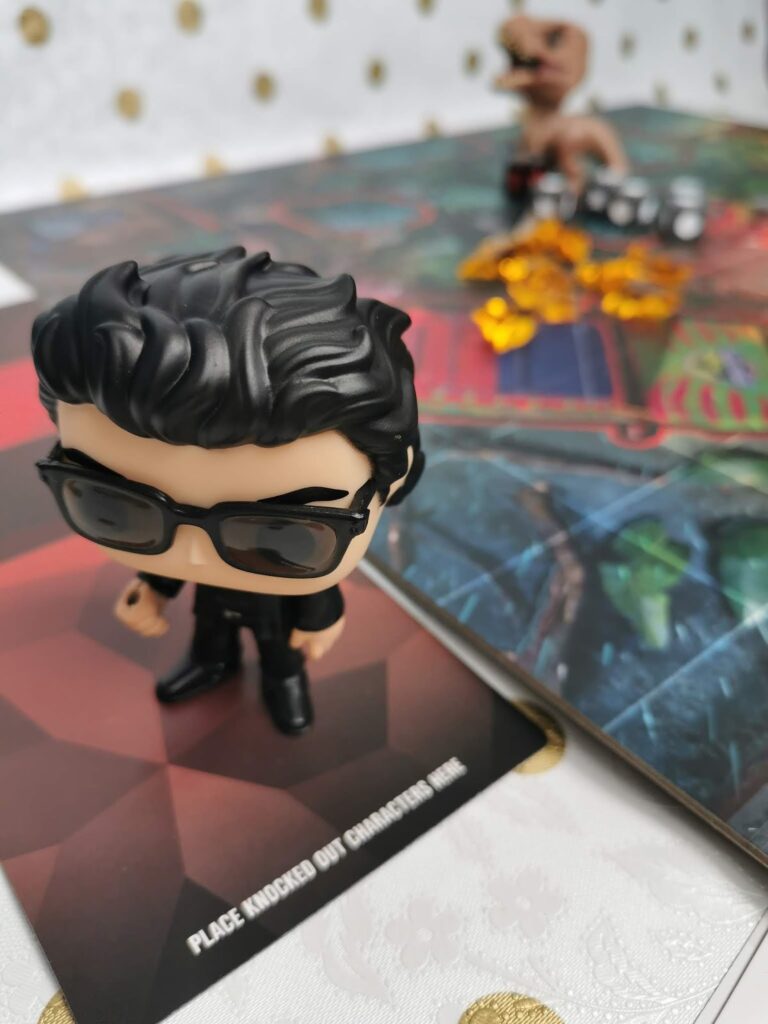 A vinyl figure of a man in black sunglasses and suit stands on a hexagonal game tile labelled "PLACE KNOCKED OUT CHARACTERS HERE." In the background, another figure and yellow game tokens sit on a colourful game board. The setting appears playful and organised.