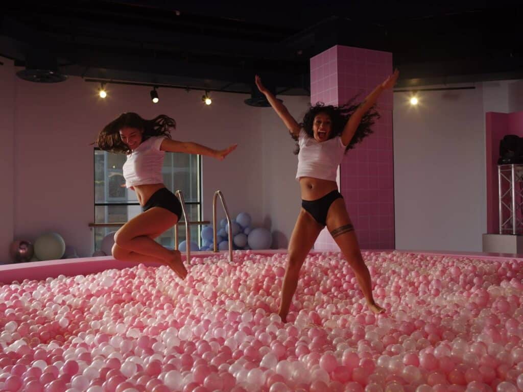 Two women in white crop tops and black bottoms joyfully jump in a large indoor pink and white ball pit. They have their arms raised in excitement. The room is brightly lit with windows, pastel decor, and spotlights on the ceiling.