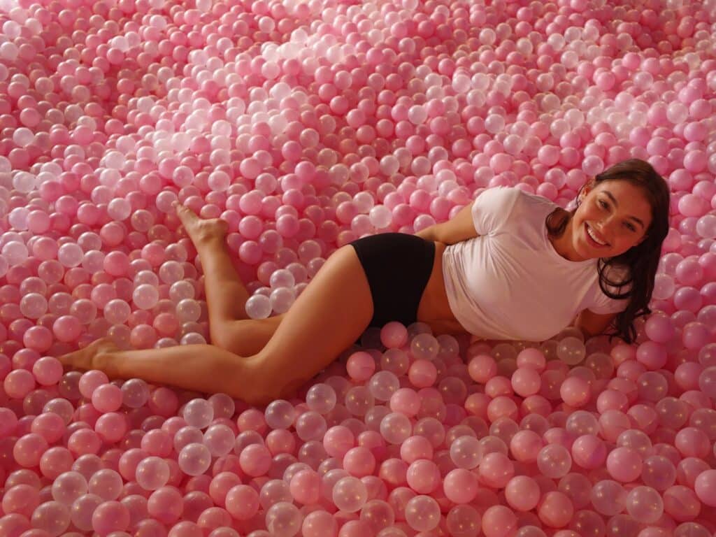 A woman in a white crop top and black shorts smiles whilst lying on her side in a large pool filled with pink and translucent plastic balls. The setting is bright, cheerful, and playful.
