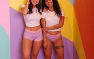 Two women stand barefoot in a colourful room with geometric pastel walls. Both wear white crop tops and striped pastel shorts. One woman has straight brown hair and touches her face, while the other has curly hair and a thigh tattoo. They are smiling and relaxed.