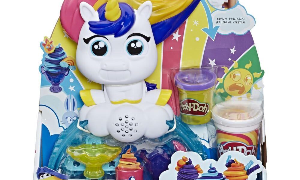 Play-Doh Tootie the Unicorn playset in colourful packaging. Tootie has a rainbow mane, large eyes, and a cheerful expression. Included are three Play-Doh pots and accessories. The background shows rainbows, stars, and Play-Doh creations. For ages 3 and over.