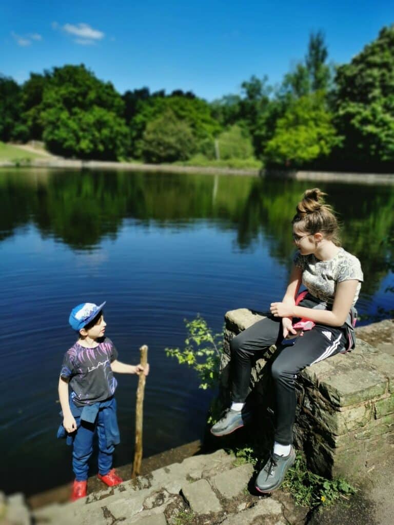 Two children by a calm lake on a sunny day. One boy in a blue cap, dark shirt, and red trainers stands on rocks holding a stick. A girl in black trousers and a patterned shirt sits on a stone wall, looking at the boy. Lush trees and blue sky are in the background.