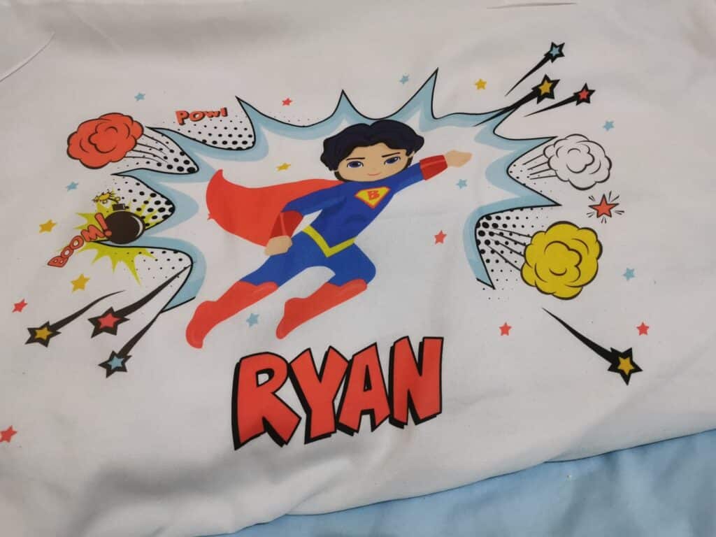 A cartoon superhero boy with dark hair, a blue suit, red cape, and "R" on his chest flies with a fist forwards. Colourful comic-style effects—"POW!", stars, and bursts—surround him. The name "RYAN" appears in bold, red, comic lettering below.