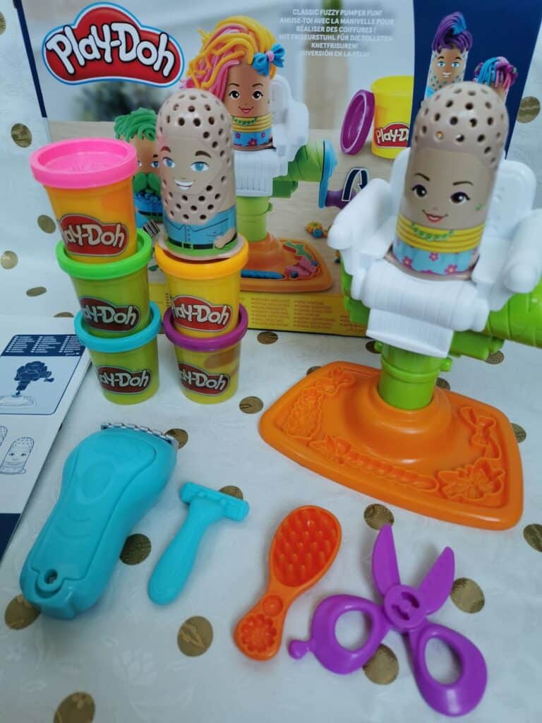 A Play-Doh hair salon playset is displayed with two doll heads in chairs, colourful Play-Doh cans stacked nearby, and toy salon tools including scissors, a brush, a comb, and a razor. The box and instruction sheet are also visible in the background.