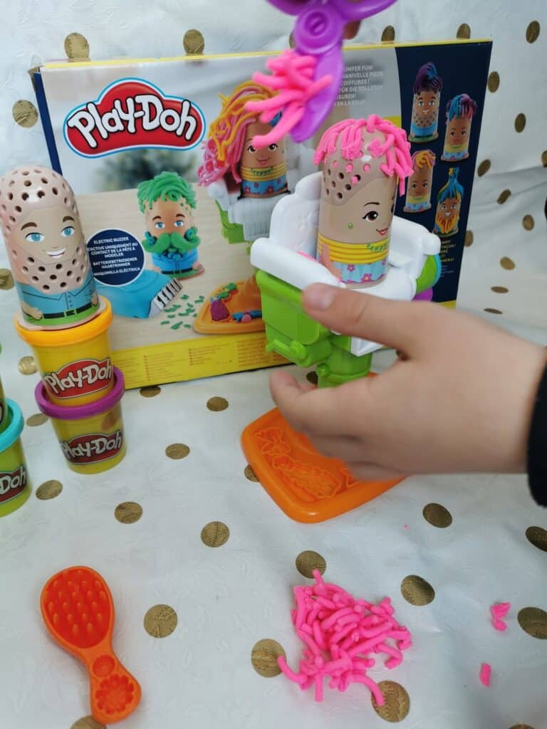 A child’s hand plays with a Play-Doh hair salon set, using a chair tool to squeeze pink Play-Doh “hair” onto a toy figure. Other Play-Doh tubs, accessories, and two more figures with Play-Doh hair are on a white table; the set box is in the background.