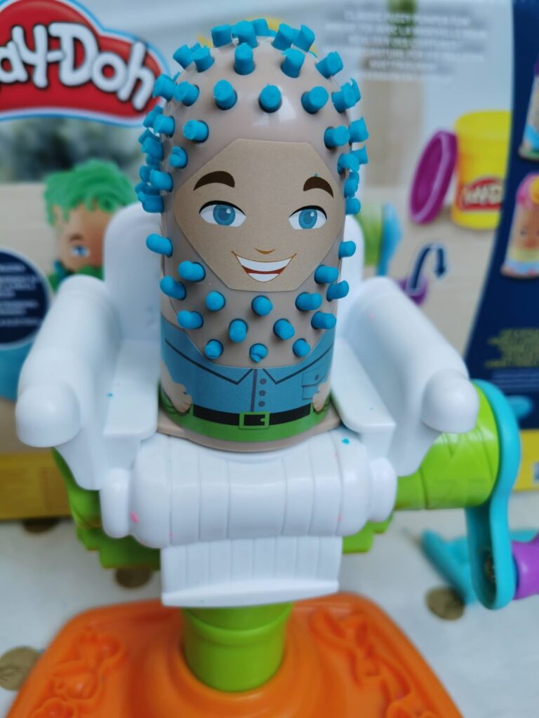 A Play-Doh toy figure with a plastic face and cartoon features sits in a white barber’s chair. Blue Play-Doh forms a beard-like texture of short spikes on its chin and head. Colourful packaging and Play-Doh containers are visible in the background.
