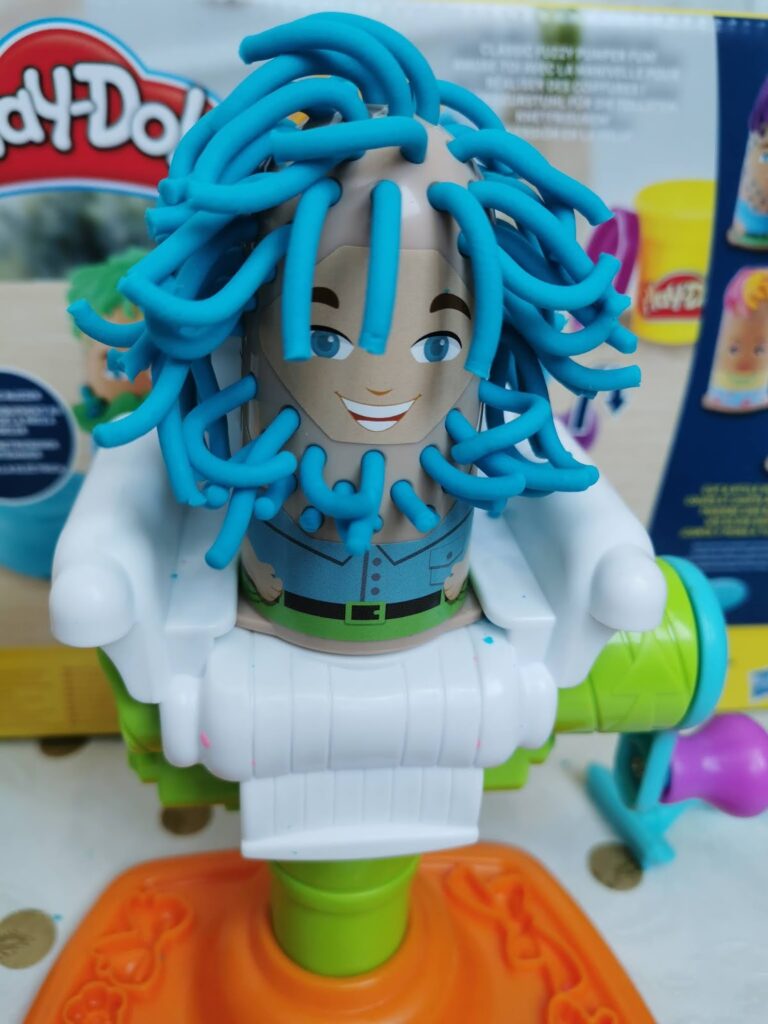 A Play-Doh toy figure with a painted-on smiling face and blue eyes sits in a white plastic chair. Blue Play-Doh strands cover its head like wild hair. The toy is part of a Play-Doh hair salon set, with colourful packaging visible in the background.
