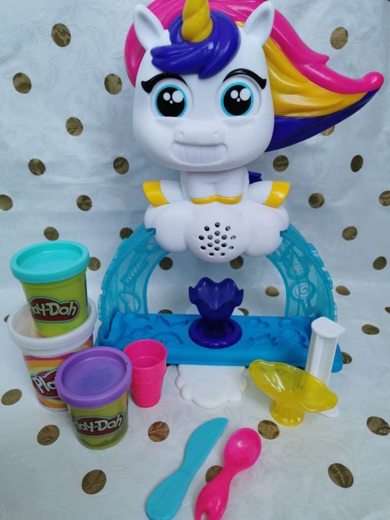 A colourful unicorn Play-Doh toy set with a white unicorn head, rainbow mane, and horn. Surrounding it are Play-Doh tubs, pink cups, blue and pink utensils, a rolling pin, and a yellow mould, all on a polka-dot background.