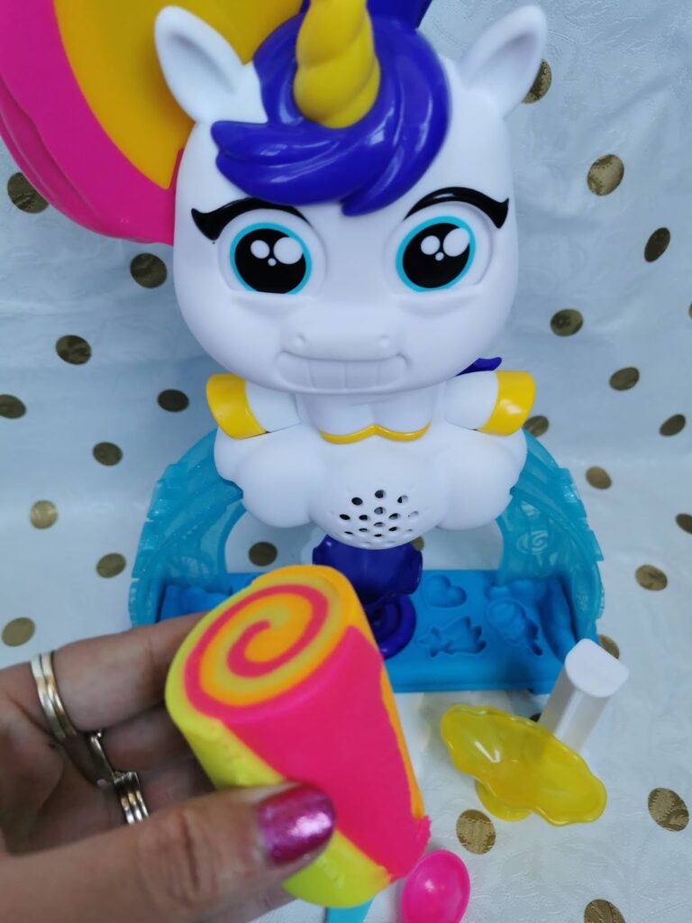 A toy unicorn with a yellow horn, blue mane, and big blue eyes sits on a blue base. In the foreground, a hand with rings holds a swirled sponge of yellow and pink. Other toy accessories, including a yellow star, are nearby. The background is white with gold dots.